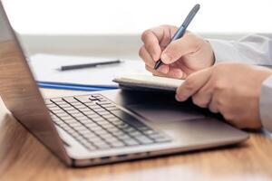 Business man write on notebook paper and use laptop computer working and analyzing with document data document for new project of business strategy at workplace. photo