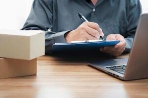 Postal man check order list for transport parcels and use laptop check stock order online order and write on the paper file photo