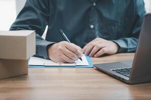 Postal man check order list for transport parcels and use laptop check stock order online order and write on the paper file photo