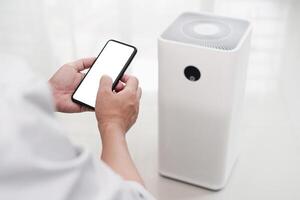 Close up hand a man holding smartphone with white screen display and online connect to air purifier using technology to control health equipment anywhere via the Internet photo