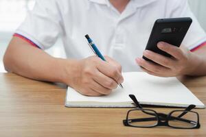 Asia man use pencil writing on notebook paper and looking smart phone with glasses on the wood table photo