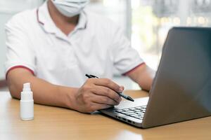 Work from home concept  Asia man wear a mask use laptop computer Find out about the spread of disease and use an clean and anti-virus alcohol spray on the wood table photo