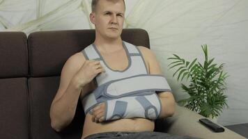 Painful, bored man with a broken arm wearing arm brace sitting on a sofa video