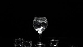 Add ice cube to shot of vodka in glass against black background. Alcohol drink video