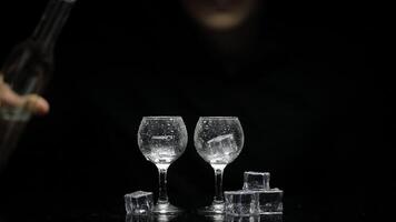 Barman pour frozen vodka from bottle into two glasses with ice. Black background video