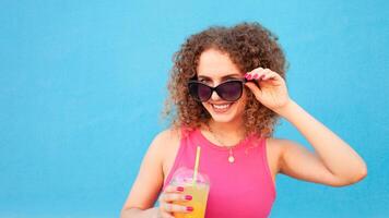 Beautiful happy woman with orange cold juice, concept summer drinks, rest and vacation. Blue background, isolated, copy space. Smiling, enjoying life, healthy and refreshing smoothie. black glasses. photo