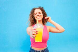 Beautiful happy woman with orange cold juice, concept summer drinks, rest and vacation. Blue background, isolated, copy space. Smiling, enjoying life, healthy and refreshing smoothie. holds out glass. photo