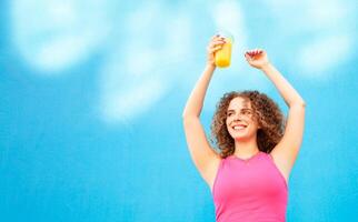 Beautiful happy woman with orange cold juice, concept summer drinks, rest and vacation. Blue background, isolated, copy space. Smiling, enjoying life, healthy and refreshing smoothie. Hands up. photo