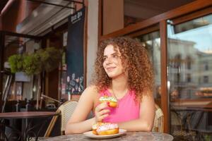 Portrait of beautiful curly young woman in cafe on summer terrace, delicious colorful donuts, sweet pastries. Lunch break, dreamy and happy. looks to left. photo