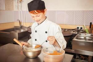 red-haired boy in chef's costume cooks pancakes in kitchen, kneads dough, assistant makes breakfast. Adds cocoa photo