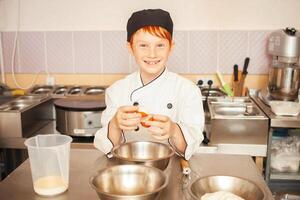 red-haired boy in chef's costume cooks pancakes in kitchen, kneads dough, assistant makes breakfast. Funny, breaks egg into bowl. photo