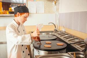 boy cooks pancakes on an industrial professional pancake maker, kitchen in cafe, little chef. Pours dough on stove photo