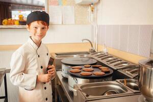 boy cooks pancakes on an industrial professional pancake maker, kitchen in cafe, little chef. red-haired child in cook's costume photo