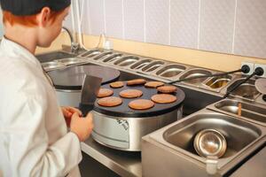 chico cocineros panqueques en un industrial profesional tortita fabricante, cocina en cafetería, pequeño cocinero. voltea panqueques con espátula foto