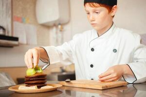 niño chico en del chef disfraz prepara chocolate panqueques en cocina en cafetería. decora panqueques con kiwi foto