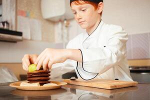 child boy in chef's costume prepares chocolate pancakes in kitchen in cafe. serious photo