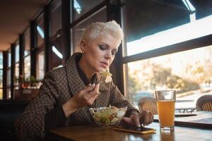 mayor mujer de 50 años es hermosa y elegante, se sienta en cafetería, trabajos con computadora portátil, Lanza libre diseñador. mira excelente, contento vida, activo estilo de vida. almuerzo, bocadillo. foto