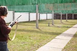 A man shoots at metal flags, targets. Firearms pump-action shotgun. Tyre's outdoor. Patrons with a fraction. Green grass, a man with headphones, a glade with protection for shooting. photo