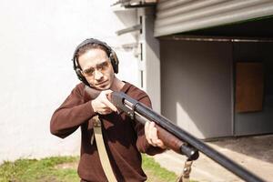 un hombre con un pomposo arma de fuego en un blanco antecedentes. un hombre en protector lentes y auriculares. neumático exterior. gris hangar. pistola foto