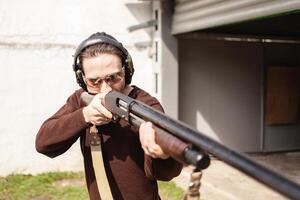 A man with a pompous firearm on a white background. A man in protective glasses and headphones. Tyre outdoor. Grey hangar. gun photo