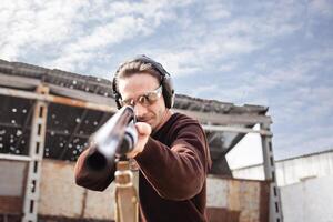 A man in protective glasses and headphones. A pump-action shotgun. Tyre's outdoors. A wall and a roof with bullet holes. Sport close up photo