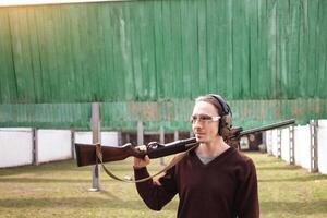 A man in protective glasses and headphones. A pump-action firearm shotgun. Tyre's outdoors. Green grass, protection for sports shooting. hobby. copy space photo