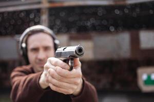 A man shoots a gun, aiming at the target. A man wearing protective headphones. A wall and a roof with bullet holes. outdoor shooting range photo