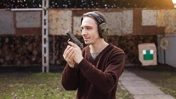 A man shoots a gun, aiming at the target. A man wearing protective headphones. A wall and a roof with bullet holes. outdoor shooting range photo