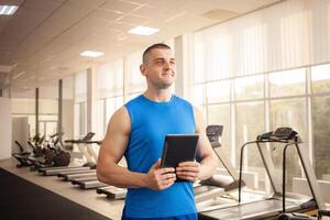 un joven hermoso entrenador en proceso de trabajando en el gimnasia. Deportes hombre con un tableta. entrenamiento, individual capacitación, físico esfuerzo, muscular cuerpo, contento sonrisas foto