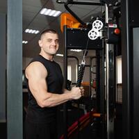 young handsome coach in a black uniform against background of a simulator in gym. Muscular athletic body of a bodybuilder, coaching, individual sports and weight loss courses. Portrait. photo