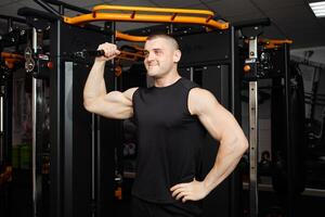 young handsome coach in a black uniform against background of a simulator in gym. Muscular athletic body of a bodybuilder, coaching, individual sports and weight loss courses. Portrait. Smiling photo