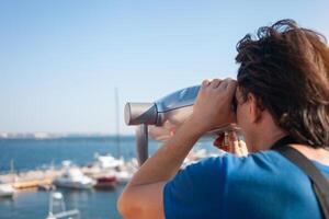 man looks through stationary binoculars. Binoscope on observation deck near sea. seaport in Odessa. Expensive yachts on the pier. Commercial machine, vending business. traveler photo