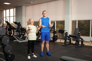 An old woman and a young male coach do exercises in gym. They shake their muscles with dumbbells, smile, are happy. Active, healthy, sporty lifestyle, senior concept indoor. photo
