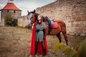 Beautiful Princess with Red Cape stands next to the horse against the backdrop of a tower and a stone wall photo