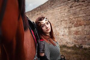 un hermosa niña soportes siguiente a un caballo en contra un Roca pared. un mujer vestido como un guerrero reina. foto