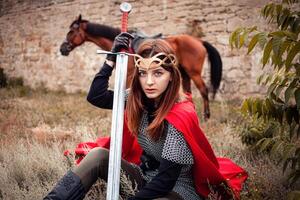 un niña con un espada en su mano en contra el antecedentes de el caballo. un mujer vestido como un guerrero reina foto