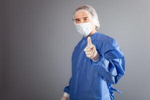A handsome male surgeon is a doctor on a gray background in the studio. A man in a robe, a medical mask, a hat. The person points his finger at the camera, calls to join something or refuse. photo