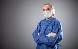 un hermoso masculino cirujano es un médico en un gris antecedentes en el estudio. un hombre en un túnica, un médico mascarilla, un sombrero. vacío espacio para texto. un hombre soportes con su brazos doblada en su pecho. foto