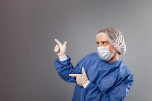 un hermoso masculino cirujano es un médico en un gris antecedentes en el estudio. un hombre en un túnica, un médico mascarilla, un sombrero. vacío espacio para texto. un hombre puntos su manos a alguna cosa a su bien. foto