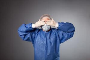 A handsome male surgeon is a doctor on a gray background in the studio. A man in a robe, a medical mask, a hat. Empty space for text. A man covers his eyes with his hands, does not want to see. photo