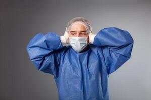 A handsome male surgeon is a doctor on a gray background in the studio. A man in a robe, a medical mask, a hat. Empty space for text. A man covers his ears, doesn't want to hear anything. photo