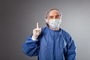 A handsome male surgeon is a doctor on a gray background in the studio. A man in a robe, a medical mask, a hat. Empty space for text. The man points his finger up, approves. photo