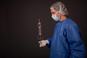 A male doctor with a terrible old syringe in his hands. The doctor in a medical mask, hat, robe and gloves. A terrible. The doctor scares with a huge syringe with a thick needle. photo