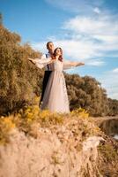 un joven Boda Pareja en amor en el borde de un acantilado en contra el fondo de el río y el cielo. un mujer en un hermosa gris vestido, un hombre en un traje en el apuntalar de el lago. foto