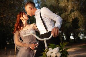 A beautiful couple in the woods on a white bike with a bouquet of flowers. Wedding walk, a woman in a gray dress, a man in a suit and suspenders looking at each other photo