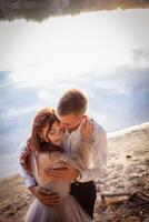 A beautiful wedding couple on the riverbank at sunset. A woman in a gray dress with flowers, a man in a suit and suspenders hug each other, love. Lovers on a walk photo