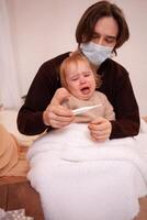 The child at home is sick, screaming and suffering. Dad in a mask checks his daughter's temperature. A child got sick, coronavirus, covid, cold, flu. A girl on the background of pills, quarantine photo