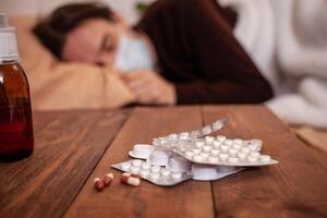 A sick man in a mask against a background of pills and medicines. The man is being treated at home in quarantine. Coronavirus, covid, cold, flu. A man fell ill in a home photo