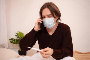 un enfermo hombre llamadas un ambulancia en el teléfono. un enmascarado hombre cheques el temperatura y diales el del doctor número en el teléfono. coronavirus, hogar cuarentena. foto