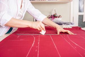 costurera a trabajar. modista haciendo ropa en moderno estudio. Sastre participación lápiz y calificación tela. mujer en pie a mesa con cortar textil, de coser máquina, hilo, patas, agujas, cinta, recortes foto